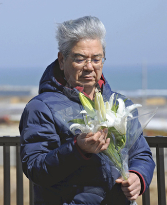 亡き妻久子さんが好きだったカサブランカを手に自宅付近に立つ山野辺清さん。「きょうの海は穏やかだよ」とカサブランカに語り掛ける