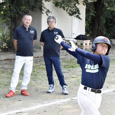 企業のステッカーをつけたユニホームやヘルメットをつけて練習する生徒を見守る佐々木さん（左）ら